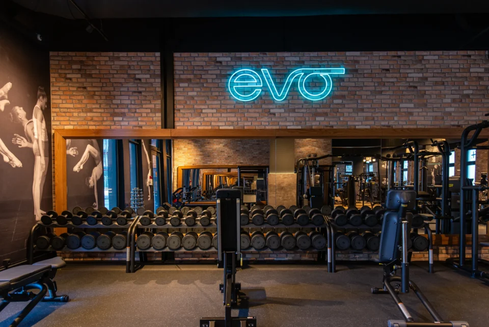 Dumbbell training zone at EVO Fitness Modecenterstraße with neon logo, exposed brick wall, and full-length mirrors