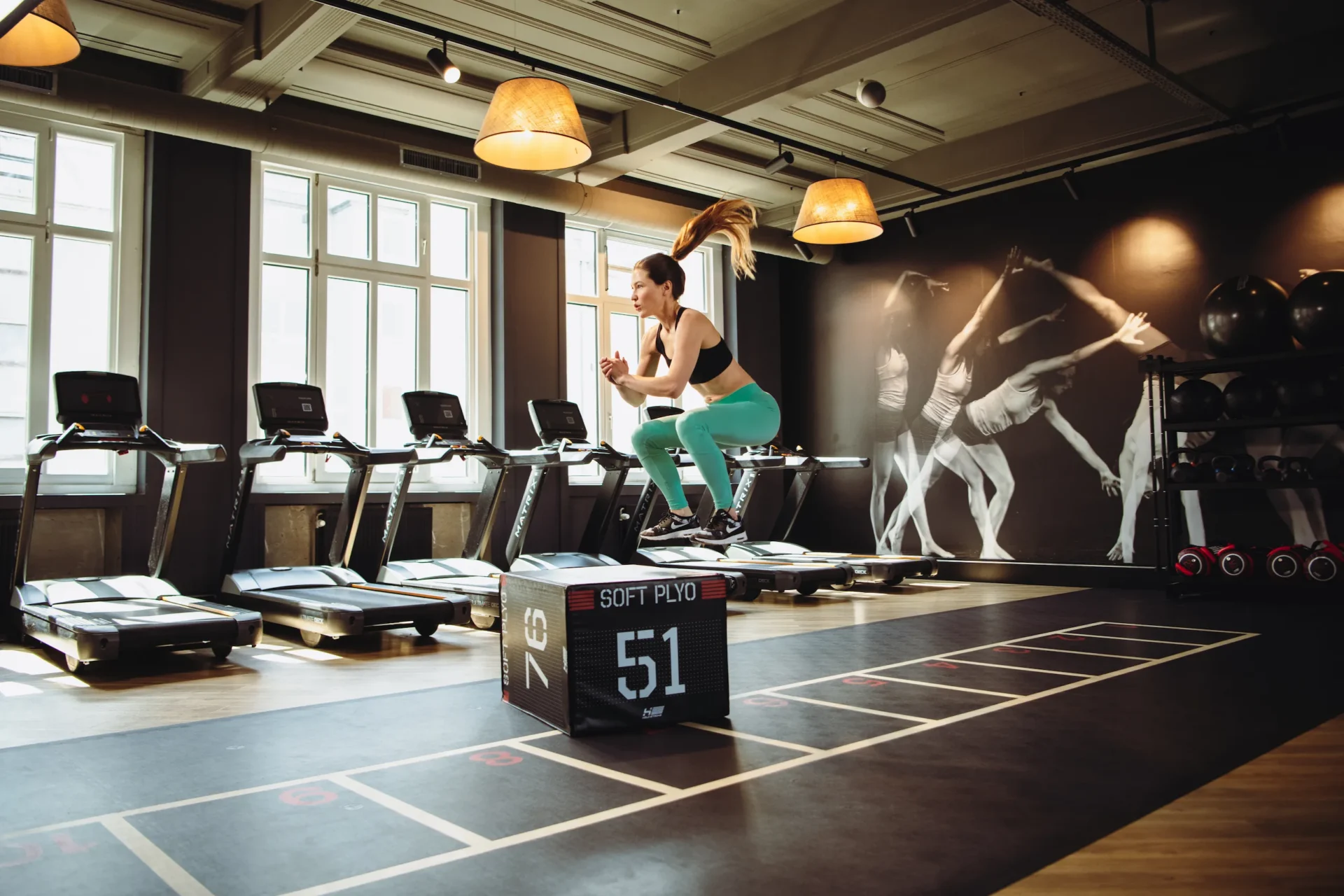 Woman mid-jump on a plyo box in front of treadmills at EVO Fitness gym