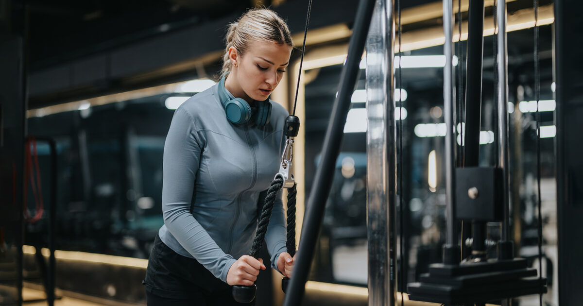 The best muscle groups to train together - Focused woman training at cable machine in the strength area of EVO Fitness, wearing headphones in a sleek, dimly lit gym.