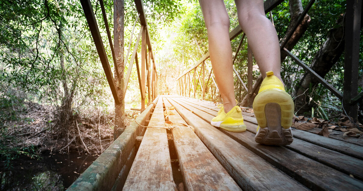 8 Wege, täglich mehr Kalorien zu verbrennen - Person mit gelben Schuhen geht ueber Holzbruecke in der Natur mit Sonne im Blick bei EVO Fitness draussen
