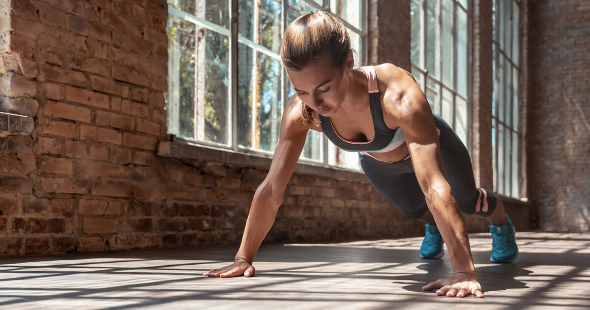 Fit on the fly: bodyweight exercises for summer vacations - Woman in a focused push-up position on a sunlit floor in the functional training area of EVO Fitness, surrounded by rustic brick walls and large industrial-style windows.