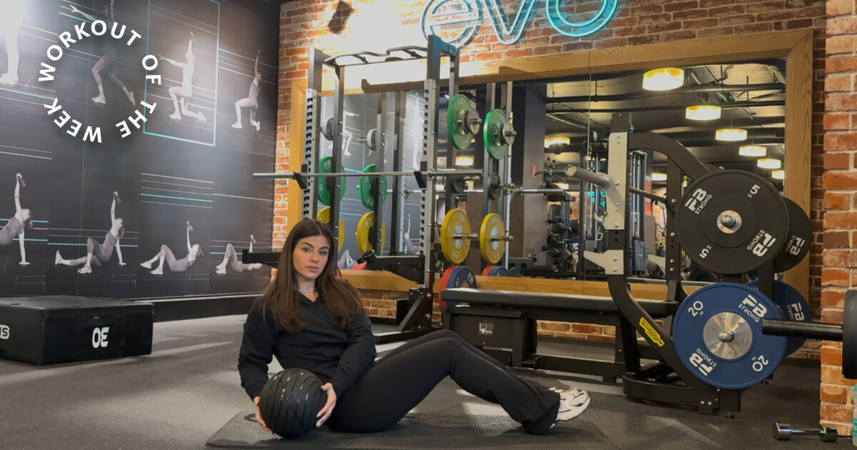 Workout of the week: 15-min core strength workout - Woman seated on mat holding medicine ball in gym with squat rack and weights behind